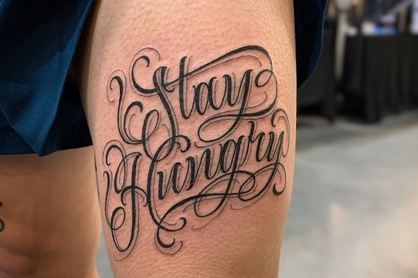 Tatuaż na udzie męski – napis  w ozdobnym stylu kaligraficznym